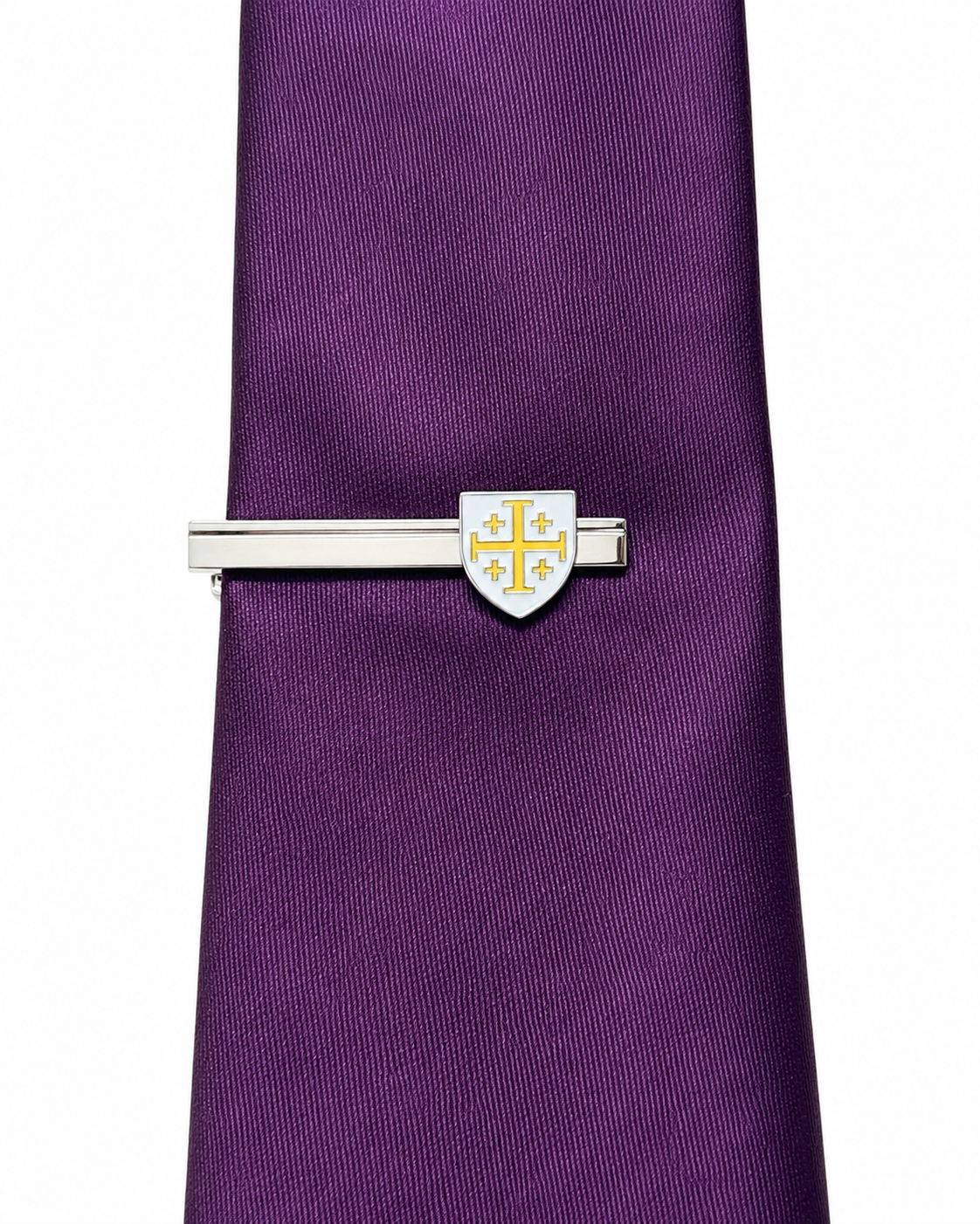 A silver-toned tie bar with a shield-shaped centerpiece featuring a gold cross, shown clipped to a purple textured necktie on a white background.