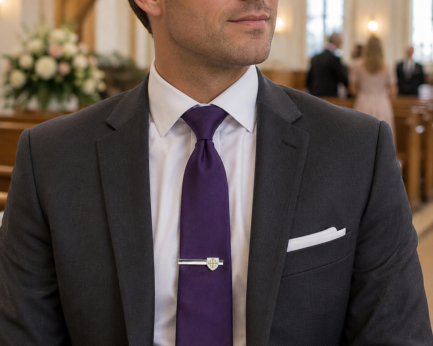 A man in a gray suit, white dress shirt, and purple tie wearing a silver-toned shield-shaped tie clip, photographed indoors with a softly blurred church or event background.