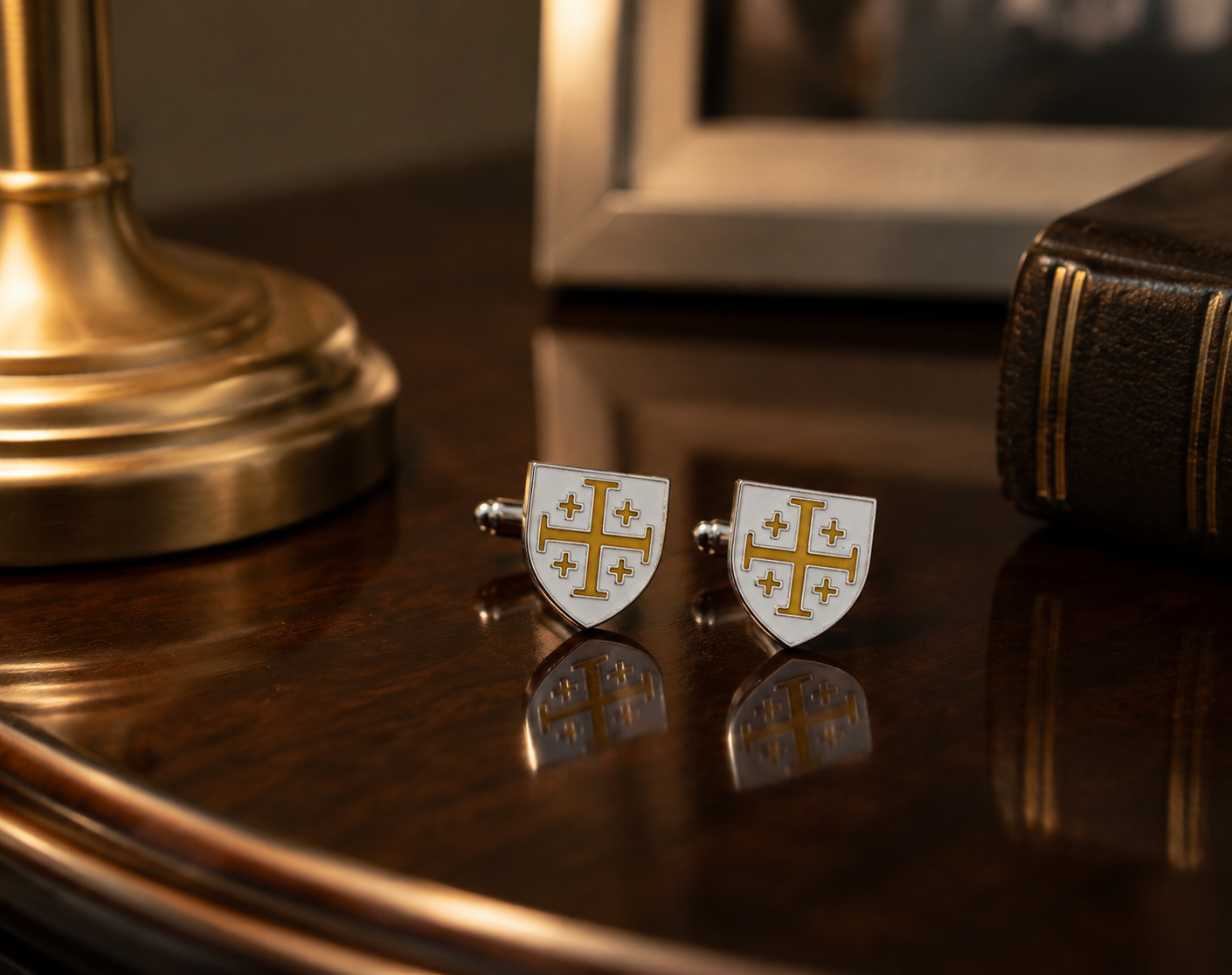 A pair of white shield-shaped cufflinks with gold crosses and stars, displayed on a polished dark wood table next to a brass lamp base and a leather-bound book.